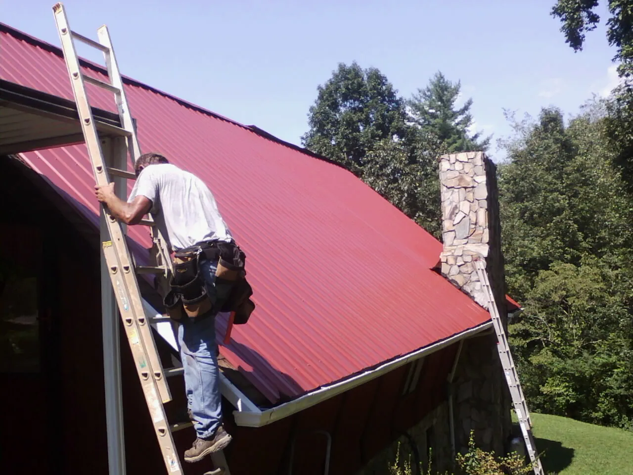 Roofer on a ladder beside a red metal roof and stone chimney — Soffit & Fascia Repair by Roofing Brothers in Pacolet